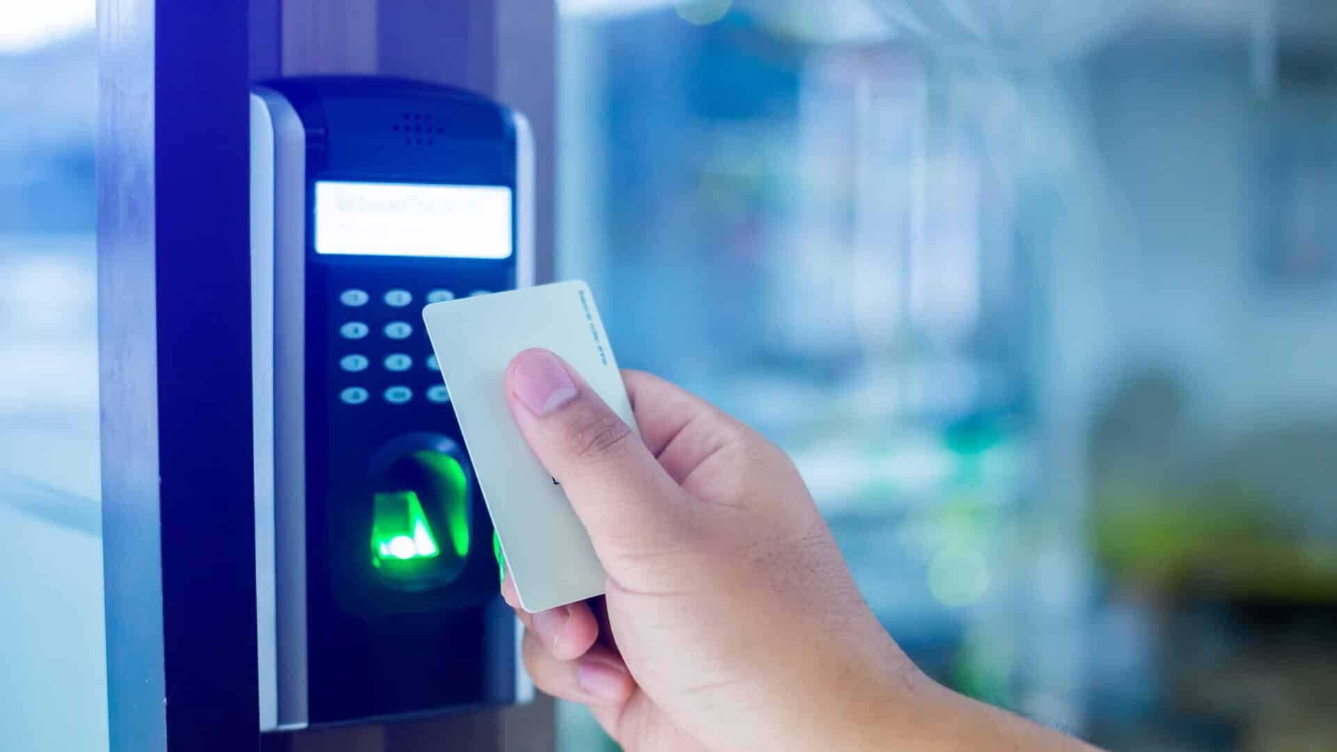 A person holds a white access card up to a card reader next to a door. The card reader has a numeric keypad and a green light indicating successful access. The background is softly blurred, suggesting an indoor environment.