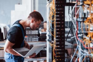 A person in work attire leans forward, holding a laptop, inspecting a server rack filled with multiple cables and equipment in a data center setting.