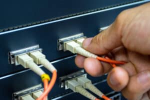 A close-up of a hand plugging an optical fiber cable into a network switch, with several other cables already connected to the device.