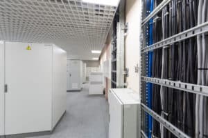 A modern server room with large white cabinets, racks of organized cables, and a grid-pattern ceiling with bright lighting.
