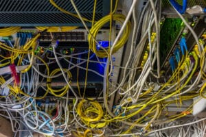 Close-up of a chaotic network server room with numerous yellow, white, and blue cables tangled and connected to various switches and hardware, some indicator lights glowing.