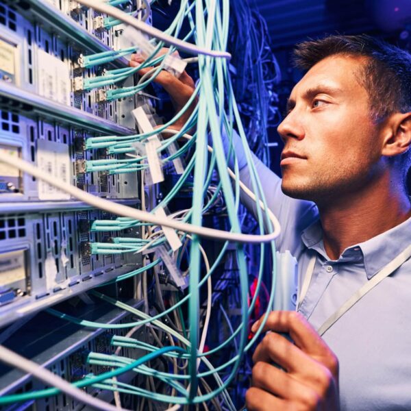 A man in a server room is connecting or inspecting blue network cables plugged into server racks, focusing closely on his work surrounded by technology equipment.
