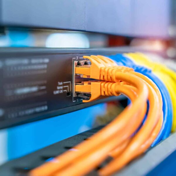 Close-up of network cables plugged into a switch, with orange and yellow cables connected to ports and out-of-focus equipment in the background, representing data networking and technology infrastructure.