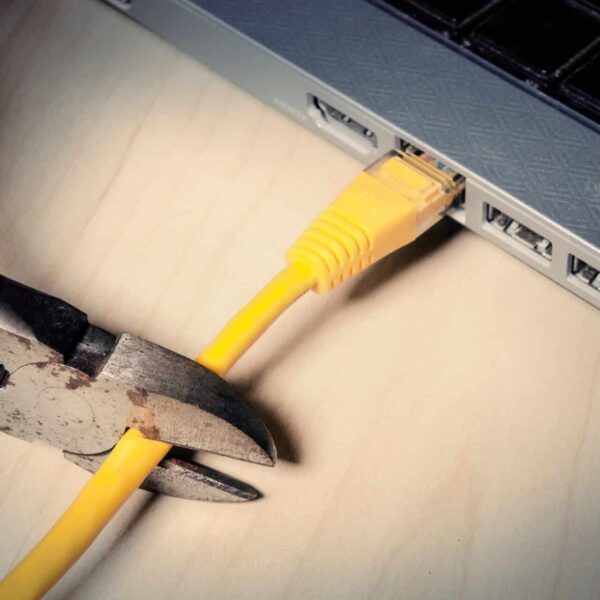 A pair of wire cutters is about to cut a yellow Ethernet cable connected to a laptop on a wooden surface.