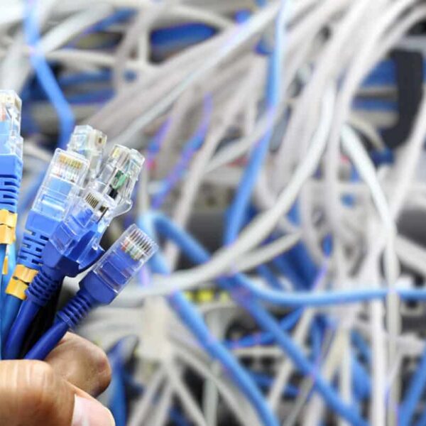 A hand holds several blue Ethernet cables in the foreground, with a tangled mess of blue and white network cables and equipment visible in the blurred background.