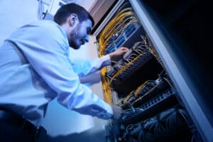 A man in a white shirt works on network cables inside a server rack, organizing and connecting wires in a data center.