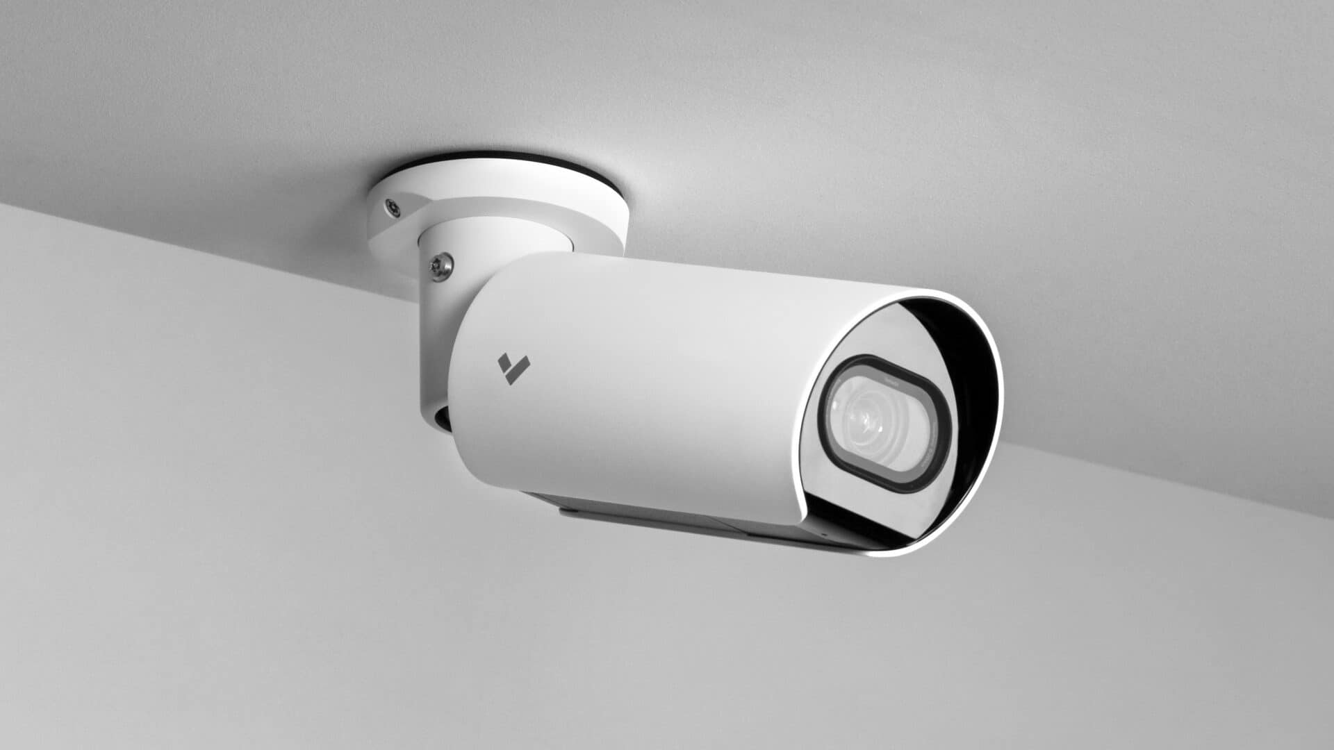 A white security camera is mounted on a ceiling, aimed outward. The background is plain and the camera has a sleek modern design.