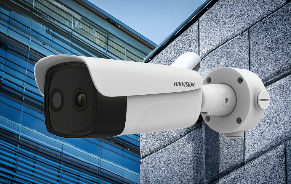 A white Hikvision security camera is mounted on a gray stone wall with a modern glass building reflected in the background.