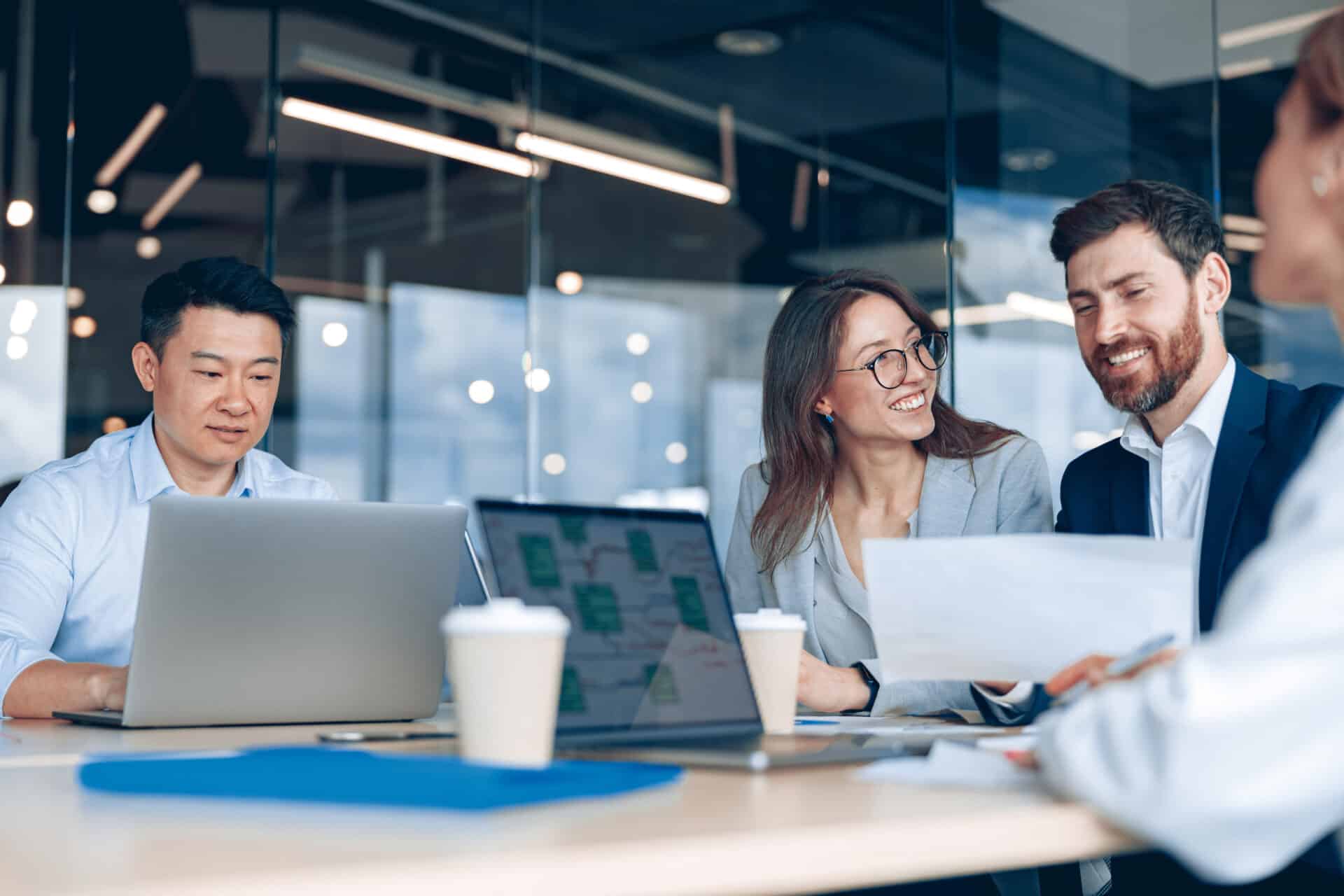 Four professionals sit at a desk in a modern office, smiling and talking while working on laptops with coffee cups and papers on the table.