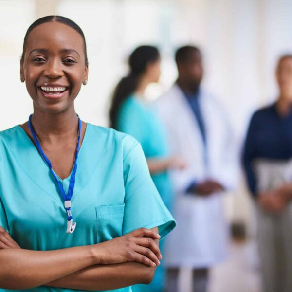 A smiling healthcare worker in teal scrubs stands with arms crossed; four colleagues converse in the bright medical background.
