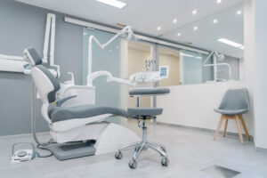 A modern dental office with a gray chair, exam tools, rolling stool, and waiting chair by a mirror wall under bright ceiling lights.