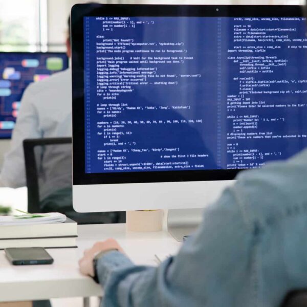 A person works at a desk with code on one monitor and diagrams on another, surrounded by books and a smartphone in a modern office.
