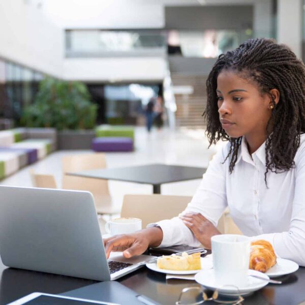 An African-American woman in a white shirt works on her laptop at a cafe table with coffee and pastries, surrounded by bright space, stairs, and plants. She is using public wi-fi.