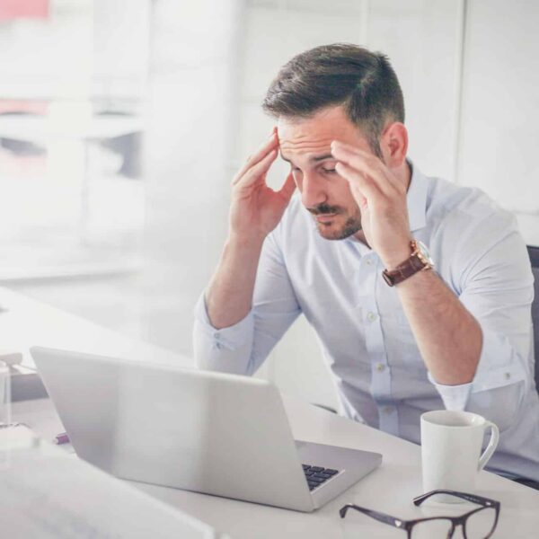 A man sits at a desk, stressed, holding his head with both hands while looking at a laptop. A coffee mug and eyeglasses are nearby.