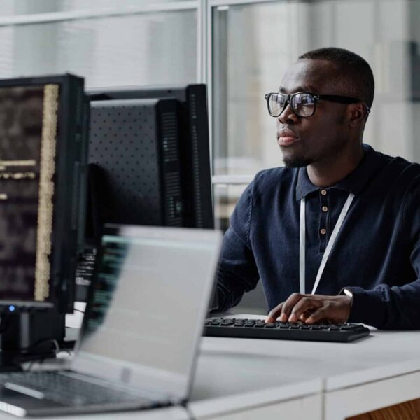 An African-American IT specialist in glasses and a dark sweater types at a desk with a monitor showing code, plus another screen and a laptop nearby.