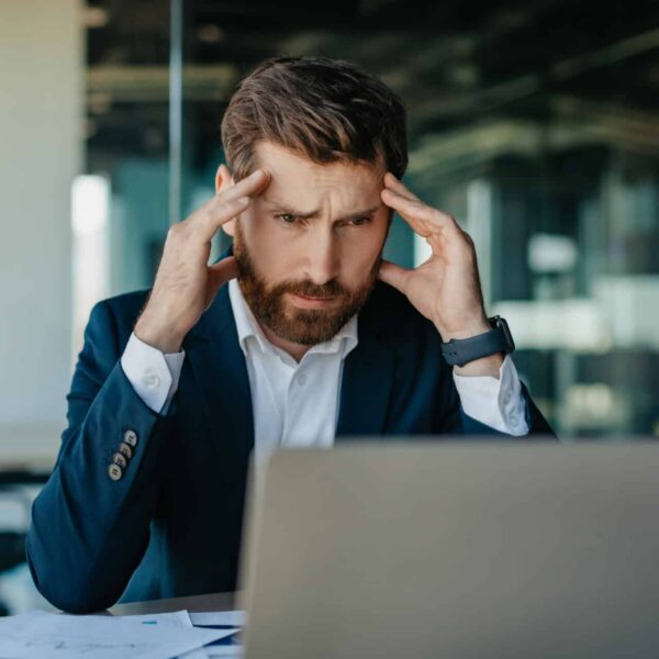 A man in a suit sits at a desk, looking stressed with both hands on his head as he stares at a laptop screen in a modern office. Indicates a cyberattack on a small business.